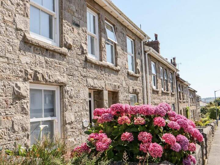 The external view of Mazey Cottage - a historic stone terraced house. There are beautiful pink flowers to the front of the house.